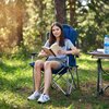 Na zdjęciu znajduje się niebieskie krzesło turystyczne NILS CAMP NC3705 ustawione na trawie w lesie. Obok krzesła stoi stół z plastikową butelką wody i książką.
