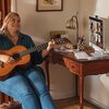 Osoba siedzi przy biurku, grając na gitarze, obok znajduje się komputer typu All-in-One z widocznym ekranem, na którym trwa wideokonferencja. Na biurku leży klawiatura, myszka oraz przybory do pisania w pojemniku.