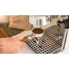 Na zdjęciu widoczny jest ekspres do kawy z metalowym portafiltrem wypełnionym zmieloną kawą, trzymanym przez rękę. W tle znajduje się worek z napisem "COFFEE" oraz metalowa tacka ociekowa z otworami.