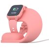 Różowa podstawka ładująca w kształcie trójkąta, na której umieszczony jest smartwatch z różowym paskiem. Z podstawki wychodzi biały kabel zasilający.