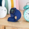 Na zdjęciu znajduje się granatowa podstawka ładująca o nowoczesnym, zaokrąglonym kształcie, obok której widnieje pudełko z napisem "STELIO JUNIOR CHARGING STAND" oraz "GRANATOWY / NAVY BLUE". Przedmiotem towarzyszy arkusz z naklejkami przedstawiającymi mo
