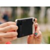 Osoba trzyma smartfon z przymocowanym czarnym powerbankiem, na którym widoczne są oznaczenia "QC", "PD MAG", "5+" oraz wskaźnik naładowania. W tle rozmyte elementy zieleni i jasne kształty.