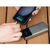 Na zdjęciu widoczny jest szary powerbank z wyświetlaczem, do którego podłączony jest smartfon z wyświetlonym poziomem naładowania 91% oraz tekstem "Szybkie ładowanie 91% 15 min do pełnego naładowania". Powerbank leży na drewnianej powierzchni, a ręce trzy
