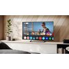 Na zdjęciu znajduje się telewizor wyświetlający ekran główny z aplikacjami takimi jak Netflix, Prime Video, YouTube, i inne. Obok telewizora stoi roślina w doniczce, a w tle widać drewnianą ścianę.