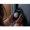 Osoba trzyma smartfon z wyświetlonym obrazem Ziemi i godziną 15:55, a na ekranie widoczny jest tekst: "Aqara Home NFC Tag". Telefon jest przyłożony do czarnego zamka szyfrowego na drzwiach.
