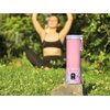 Różowy blender personalny z napisem "BlendyGo" stoi na kamieniu w otoczeniu zieleni. W tle widoczna jest osoba w stroju sportowym, siedząca na trawie.