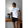 Na zdjęciu widoczny jest pojemnik z odżywką białkową "Iso Whey Zero" marki BioTechUSA, stojący na drewnianym stole. Obok stoi osoba trzymająca shaker z napisem "BioTechUSA" na koszulce.