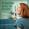 Na zdjęciu widoczna jest osoba trzymająca miętową szczoteczkę soniczną z napisem "A better way to brush" oraz "Elektryczna szczoteczka soniczna Do 80 000 wibracji/min." w tle. W tle znajduje się rozmyty wzór w paski oraz roślina.
