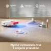 Na zdjęciu widoczny jest biały robot sprzątający na drewnianej podłodze, z fragmentem dywanu po prawej stronie. Na obrazie znajduje się tekst: "Zwiększenie mocy ssania na dywanach" oraz "Ultradźwiękowe wykrywanie dywanów".