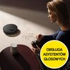 Czarny robot sprzątający znajduje się na jasnej podłodze, obok rozsypanych drobinek. Na zdjęciu widoczny jest również okrągły, żółty znak z napisem "OBSŁUGA ASYSTENTÓW GŁOSOWYCH".