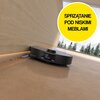 Czarny robot sprzątający znajduje się na podłodze przy ścianie, zbierając drobne zanieczyszczenia. W prawym górnym rogu widnieje żółte koło z napisem "SPRZĄTANIE POD NISKIMI MEBLAMI".