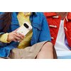 Osoba trzyma w ręku jasny smartfon z widocznym logo w kształcie litery "G". Ubrana jest w żółty sweter, niebieską dżinsową kurtkę i beżową spódnicę, a obok niej znajduje się osoba w czerwonej kurtce.