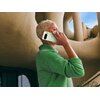 Osoba ubrana w zielony sweter trzyma przy uchu smartfon w kolorze księżycowej szarości, na tle nowoczesnej architektury z zaokrąglonymi kształtami. Na telefonie widoczne jest logo producenta.
