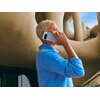 Osoba trzyma przy uchu smartfon w kolorze księżycowej szarości, ubrana w niebieski sweter. W tle widoczna jest nowoczesna architektura z zaokrąglonymi kształtami i otworami.