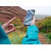 Osoba w niebieskiej kurtce trzyma smartfon z widocznym ekranem, na którym wyświetla się abstrakcyjny wzór. W tle znajduje się krajobraz z zielenią i skałami.