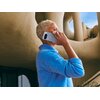 Osoba trzyma przy uchu smartfon w kolorze księżycowej szarości, na tle nowoczesnej architektury z zaokrąglonymi kształtami. Na obudowie telefonu widoczne jest logo producenta.