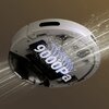 Na zdjęciu znajduje się biały robot sprzątający o okrągłym kształcie, poruszający się po powierzchni, która przechodzi z gładkiej na teksturowaną. W tle widoczne są graficzne elementy w kolorach niebieskim i pomarańczowym, symbolizujące przepływ powietrza
