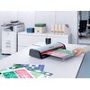 Na zdjęciu znajduje się laminator z napisem "Series 2" na biurku, z którego wychodzi zalaminowany arkusz z kolorowymi grafikami. W tle widoczne są segregatory, pudełka oraz drukarka biurowa.
