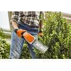 Na zdjęciu widoczne są akumulatorowe nożyce do żywopłotu w trakcie przycinania trawy. Na obudowie urządzenia znajduje się logo "STIHL" oraz kilka piktogramów ostrzegawczych.