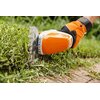 Na zdjęciu widoczne są akumulatorowe nożyce do żywopłotu z logo "STIHL" na obudowie, trzymane przez osobę w pomarańczowych rękawicach ochronnych. W tle znajduje się ogród z zielonymi krzewami i drzewami oraz fragment budynku.
