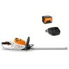 Na zdjęciu widoczne są akumulatorowe nożyce do żywopłotu w kolorach białym, pomarańczowym i czarnym, z logo "STIHL" na obudowie. Obok znajdują się akumulator z napisem "STIHL AK 10" oraz czarna ładowarka z logo "STIHL".