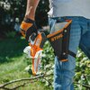 Na zdjęciu widoczna jest pomarańczowo-biała piła akumulatorowa z napisem "STIHL" trzymana w ręku osoby w rękawicach ochronnych. Obok znajduje się czarna kabura z pomarańczowym napisem "STIHL" przymocowana do niebieskich dżinsów.