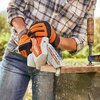 Na zdjęciu widoczna jest piła akumulatorowa z logo "STIHL" na obudowie, używana do cięcia drewnianej deski. W tle znajduje się rozmyty krajobraz, a na desce widoczne są trociny unoszące się w powietrzu.
