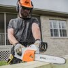 Na zdjęciu widoczna jest osoba używająca elektrycznej piły łańcuchowej marki STIHL, z widocznym logo na obudowie piły. W tle znajduje się budynek z ceglaną elewacją i dużymi oknami.