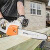 Na zdjęciu widoczna jest piła łańcuchowa elektryczna z logo "STIHL" na obudowie oraz napisami "STIHL ROLLOMATIC E" i "Made in Germany" na prowadnicy. Piła przecina drewnianą belkę, a w tle znajduje się budynek z ceglaną elewacją i oknami.