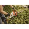 Na zdjęciu widoczne są elektryczne nożyce do żywopłotu marki STIHL HSE 52, trzymane przez osobę w rękawicach roboczych. Nożyce są używane do przycinania zielonego żywopłotu w ogrodzie, a na obudowie narzędzia widnieje logo "STIHL".