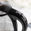 Na zdjęciu znajduje się przezroczysty czajnik elektryczny z czarnym uchwytem i metalową podstawą, ustawiony na jasnym blacie. W tle widoczne są niebieski dzbanek, filiżanki oraz słoik z orzechami.