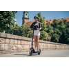Na zdjęciu widoczna jest czarna hulajnoga elektryczna z dużymi kołami i amortyzacją, użytkowana na tle kamiennego muru i zieleni. W tle znajdują się drzewa oraz fragmenty budynków z czerwonymi dachami i wieżami.