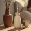 Na zdjęciu znajduje się biały aromatyzer wydzielający delikatną mgiełkę, obok którego stoi buteleczka z napisem "Stadler Form LAVENDER LAVANDULA ANGUSTIFOLIA PURE ESSENTIAL OIL". W tle widoczny jest brązowy wazon z gałązkami oraz filiżanka.