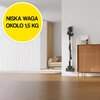 Na zdjęciu znajduje się odkurzacz przymocowany do ściany obok drewnianej szafki, a w tle widoczna jest sofa z poduszkami. W lewym górnym rogu znajduje się żółte koło z napisem "NISKA WAGA OKOŁO 1,5 KG".