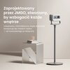 Na zdjęciu znajduje się stojak z zamontowanym urządzeniem, obok którego stoi nowoczesny stolik z książkami i szklanym przedmiotem na blacie. Na ścianie widoczny jest tekst: "Zaprojektowany przez JMGO, stworzony, by wzbogacić każde wnętrze. Uniwersalny, mi