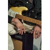 Na zdjęciu widoczna jest gitara elektryczna w jasnym kolorze, oparta na kolanach osoby siedzącej, która ma na palcach kilka pierścieni. Obok znajduje się wzmacniacz z widocznym logo "Laney".