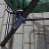 Zbliżenie na metalową ramę ogrodowego fotela z zielonym materiałem, stojącego na szarej, brukowanej powierzchni. Widoczny jest mechanizm łączenia elementów ramy z dwoma śrubami.