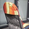 Na zdjęciu widoczny jest fragment ogrodowego fotela z kolorowym, wzorzystym obiciem w odcieniach pomarańczowego, żółtego i czerwonego. W tle znajdują się przeszklone drzwi z metalową klamką.