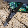 Fragment fotela ogrodowego z czarną metalową ramą i kolorowym materiałem w zielone liście, ustawiony na podłożu z jasnych kamieni. Widoczny jest mechanizm składania fotela.