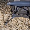 Na zdjęciu widoczny jest fragment leżaka ogrodowego z metalową ramą i materiałowym siedziskiem, ustawiony na podłożu z drobnych kamieni. Konstrukcja leżaka jest w kolorze ciemnym, a na jednym z elementów znajduje się mała, okrągła naklejka.