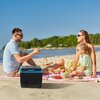 Na zdjęciu widać lodówkę kompresorową z napisem "Webber" na plaży, obok której znajduje się koc piknikowy z koszykiem i jedzeniem. W tle widoczna jest linia drzew oraz spokojna woda pod błękitnym niebem.