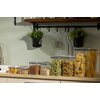 Na zdjęciu znajduje się zestaw ośmiu przezroczystych pojemników kuchennych wypełnionych różnymi produktami spożywczymi, takimi jak makarony, płatki i orzechy, ustawionych na blacie kuchennym. W tle widoczna jest ściana z wieszakiem na przybory kuchenne or
