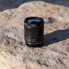Czarny obiektyw fotograficzny z widocznymi oznaczeniami "Canon 24-105" stoi na jasnym, nierównym kamieniu. Obiektyw ma wyraźne pierścienie do regulacji ostrości i zoomu.