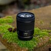 Czarny obiektyw fotograficzny z widocznym napisem "Canon 24-105" stoi na powierzchni pokrytej zielonym mchem. W tle znajdują się rozmyte elementy natury oraz szyszki.