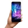 Na zdjęciu widoczna jest dłoń trzymająca smartfon z kolorowym, abstrakcyjnym tłem na ekranie. Obok telefonu znajduje się przezroczysta folia ochronna, lekko przesunięta względem ekranu.