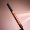 Na zdjęciu znajduje się suszarko-lokówka w kolorze bursztynowym z czarną końcówką, umieszczona na tle w odcieniach różu. Produkt ma cylindryczny kształt z widocznymi przyciskami i perforowaną częścią na dole.