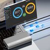 Na zdjęciu znajduje się srebrna obudowa dysku z widocznym logo "Zenwire" umieszczona na klawiaturze laptopa. W tle widoczny jest ekran z wynikami testu prędkości, pokazujący wartości "3071 MB/s" dla zapisu i "3094 MB/s" dla odczytu, a także ikony symboliz