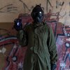 Osoba w zielonym kombinezonie i czarnej masce trzyma w ręku urządzenie z wyświetlonym symbolem tarczy. W tle widoczna jest ściana pokryta graffiti i napisami.