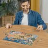 Na zdjęciu widać osobę układającą puzzle na drewnianym stole, z widocznym fragmentem układanki przedstawiającym kolorową, oceaniczną scenę. W tle znajduje się roślina doniczkowa oraz zasłona.