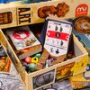 Otwarta pudełko gry planszowej "Projekt A.R.T." marki Muduko zawiera karty z różnymi symbolami, kolorowe kostki z numerami oraz drewniane elementy w kształcie skrzyń i serc. Na pudełku widoczne są ilustracje oraz logo Muduko.